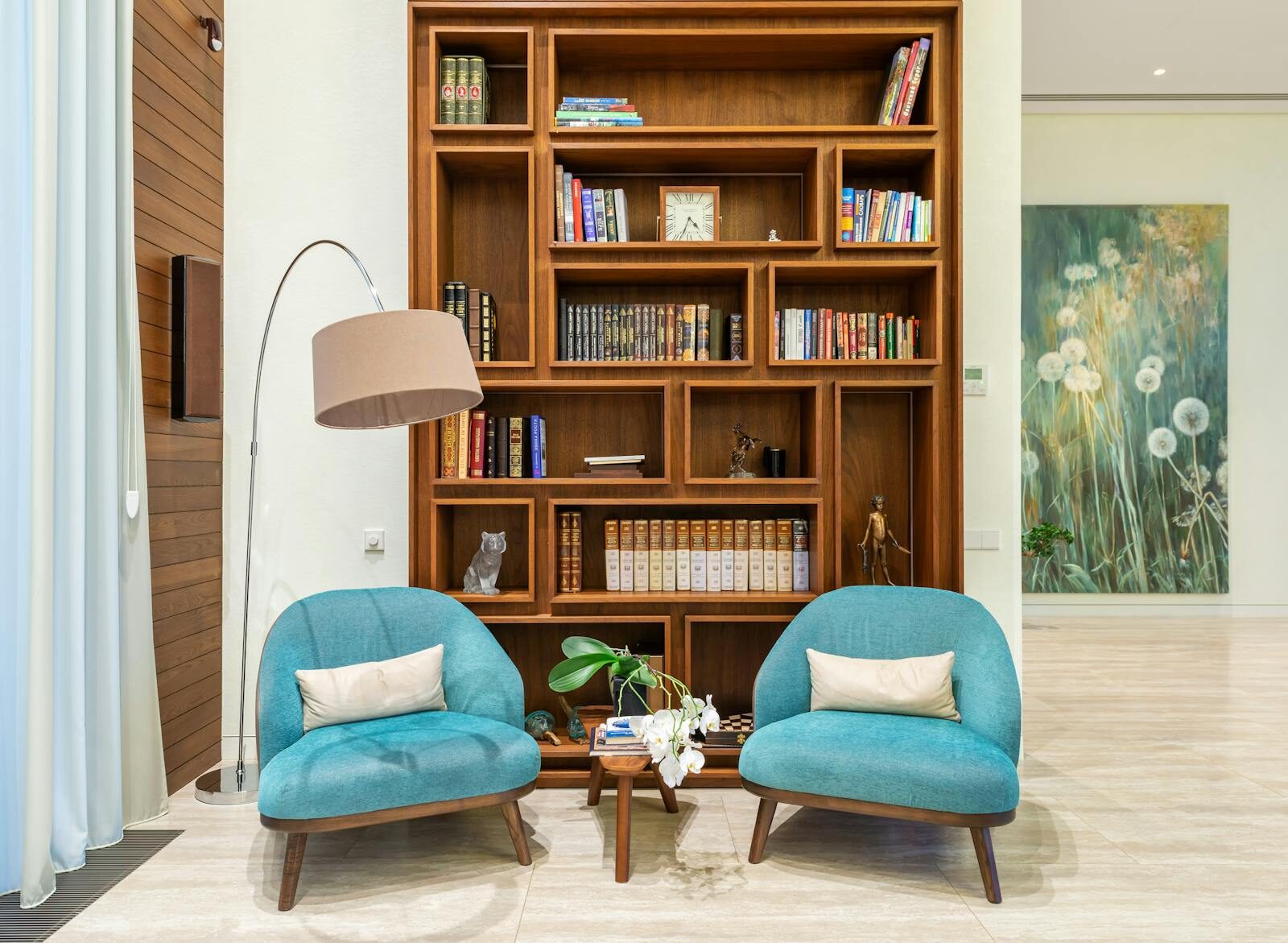Interior of living room with cozy soft blue armchairs near lamp and books placed on shelves of wooden bookcase