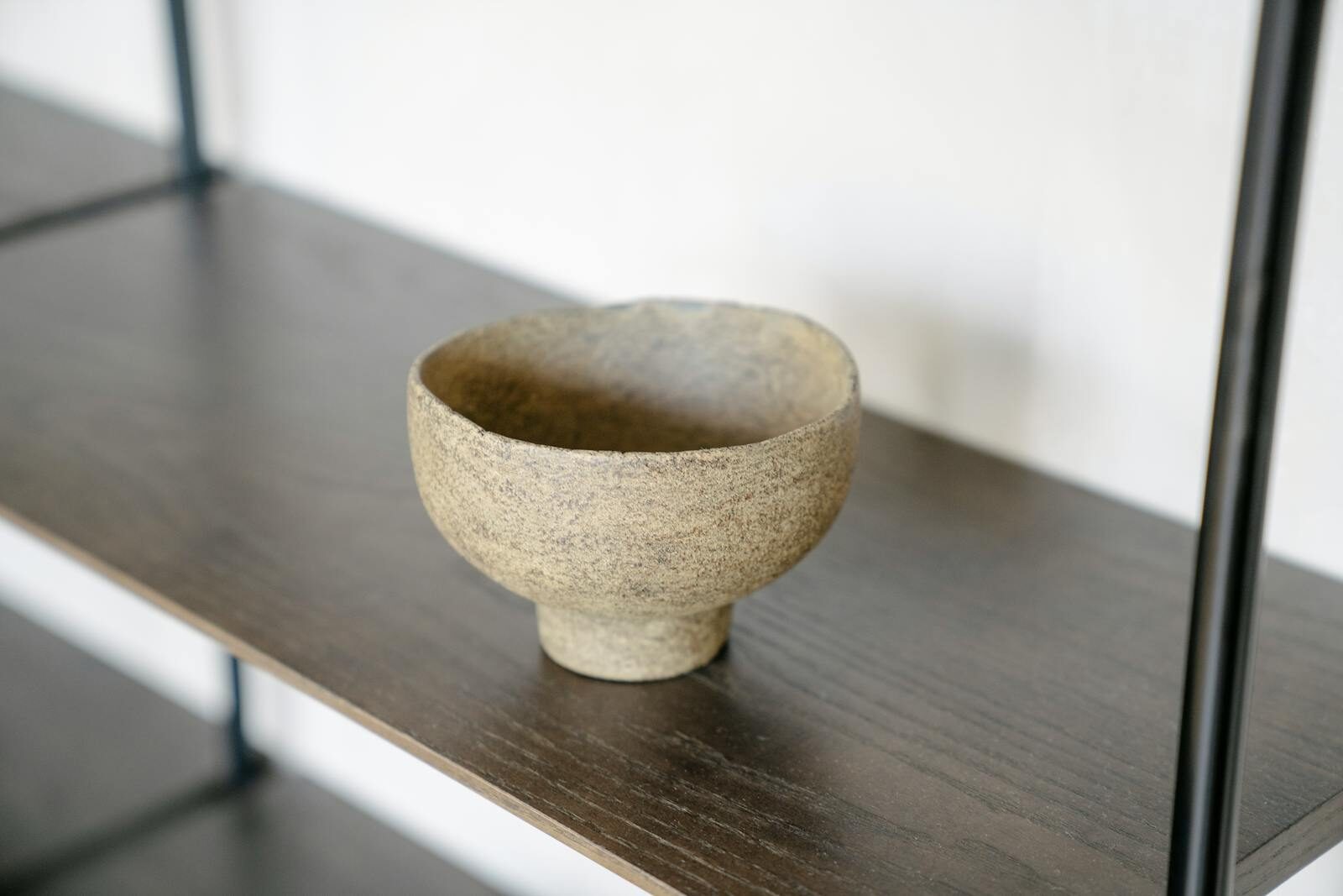 A close-up of a handmade rough clay bowl placed on a dark wooden shelf.